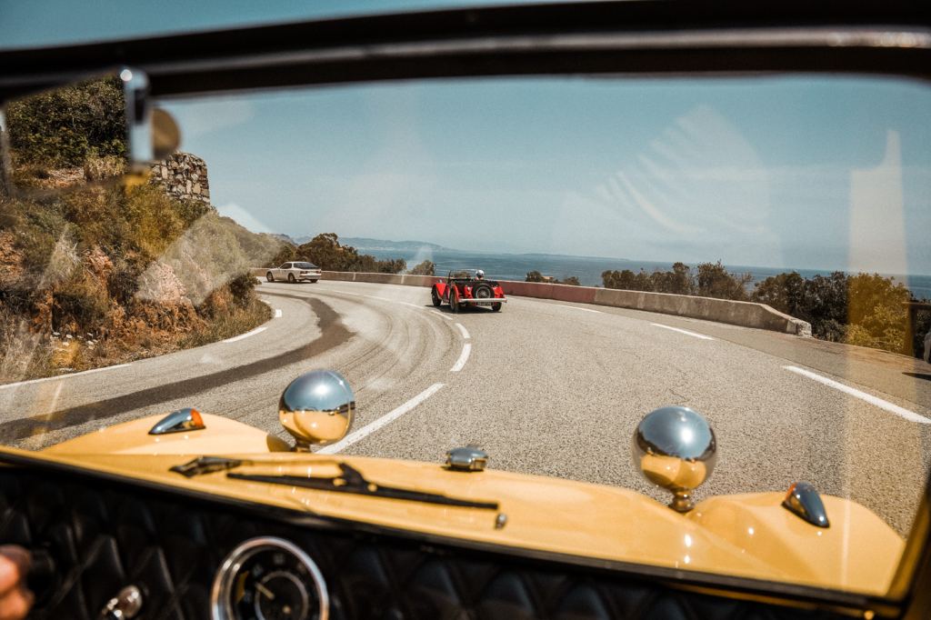 Blick aus einem gelben Oldtimer über eine Küstenstrasse, vorweg fahren zwei weitere Oldtimer, rechts liegt das Mittelmeer.