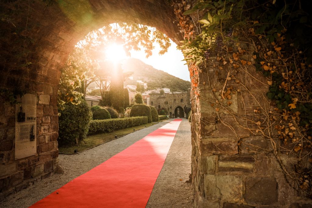 Roter Teppich führt durch einen efeubewachsenen Torbogen auf ein Château zu, die Abendsonne steht tief über dem bewaldeten Hügel.