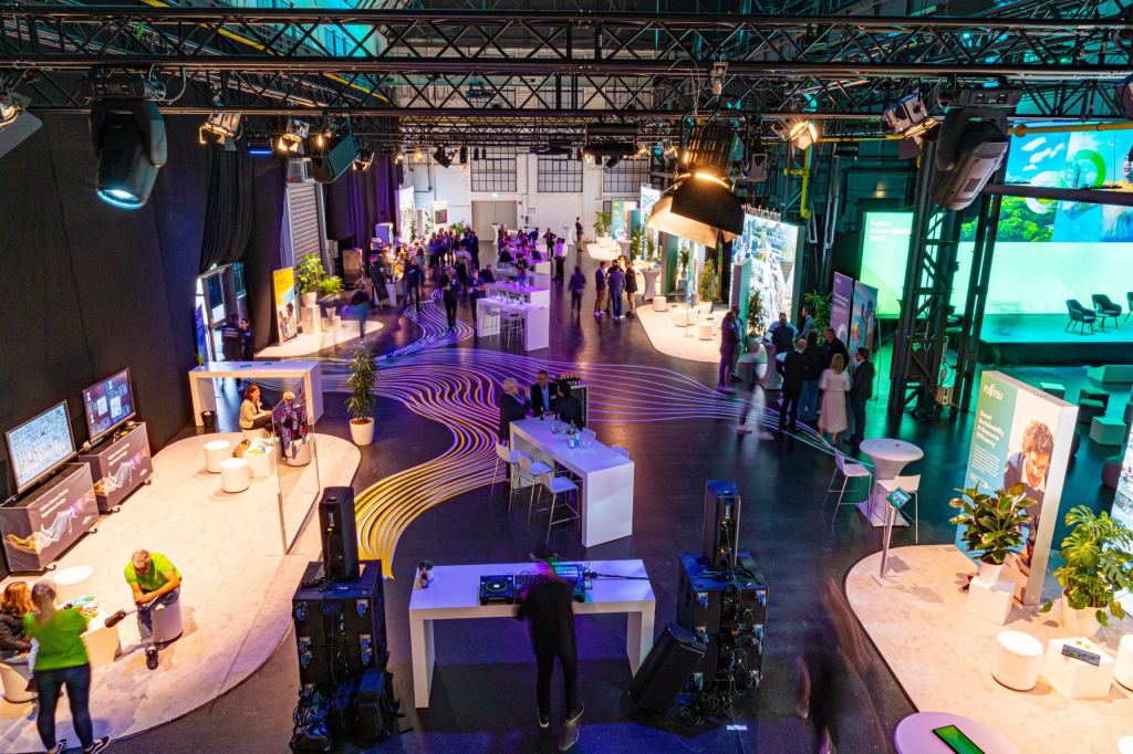 Blick von oben auf eine Fujitsu-Veranstaltung in einer Industriehalle mit geschwungenen Lichtbändern auf dem Boden, Ausstellungsinseln, Stehtischen und einem DJ-Pult.