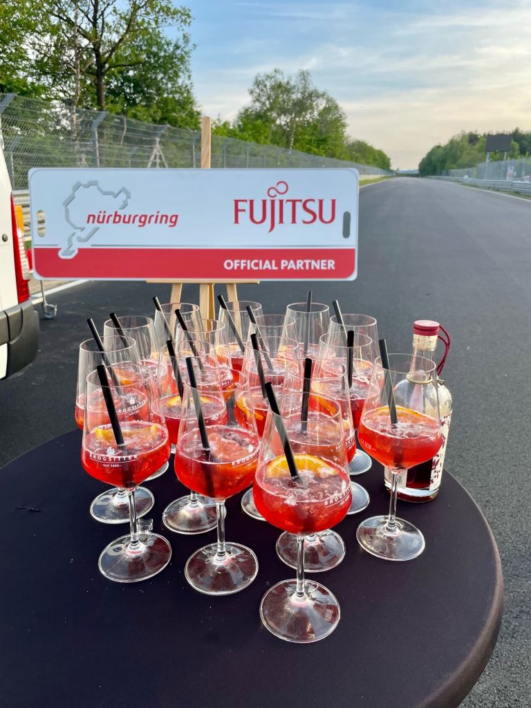 Rund zwanzig Gläser mit rotem Aperitif auf einem Stehtisch unmittelbar an der Rennstrecke, dahinter ein Schild mit den Logos von Nürburgring und Fujitsu und dem Zusatz Official Partner.