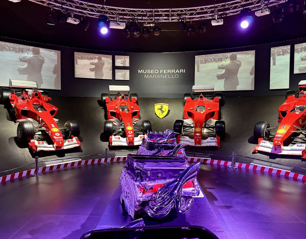 Ausstellungsraum des Museo Ferrari in Maranello mit vier roten Formel-1-Rennwagen an einer geschwungenen Wand, darüber historische Schwarzweissaufnahmen auf Bildschirmen, in der Mitte das Ferrari-Wappen und im Vordergrund ein Motorblock unter violettem Scheinwerferlicht.