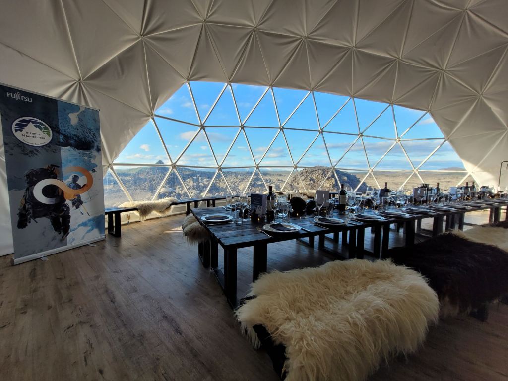 Lange eingedeckte Tafel in einer Kuppel aus Glasdreiecken mit Blick auf die isländische Lavalandschaft, auf den Bänken liegen Schaffelle.