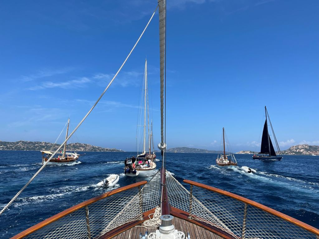 Blick vom Bug eines Segelschiffs auf vier weitere Segelyachten, die vor der felsigen Küste Sardiniens in Fahrt sind.