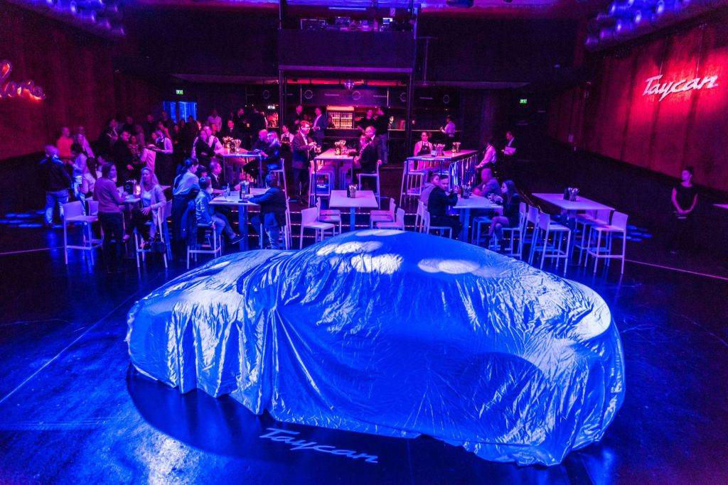 Blau ausgeleuchteter Saal vor einer Fahrzeugpremiere: Unter einer Abdeckplane mit Taycan-Schriftzug zeichnet sich ein Sportwagen ab, dahinter warten Gäste an Stehtischen.
