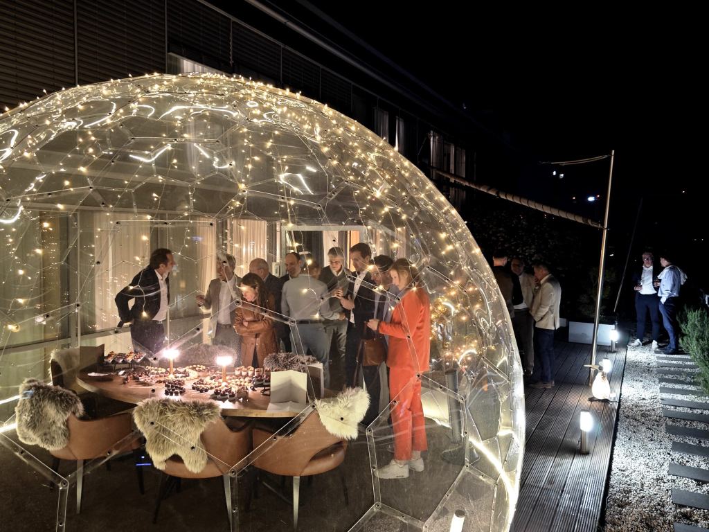 Transparente Iglu-Kuppel mit Lichterketten auf einer Terrasse, darin stehen Gäste um eine Tafel mit Desserts und Kerzen.