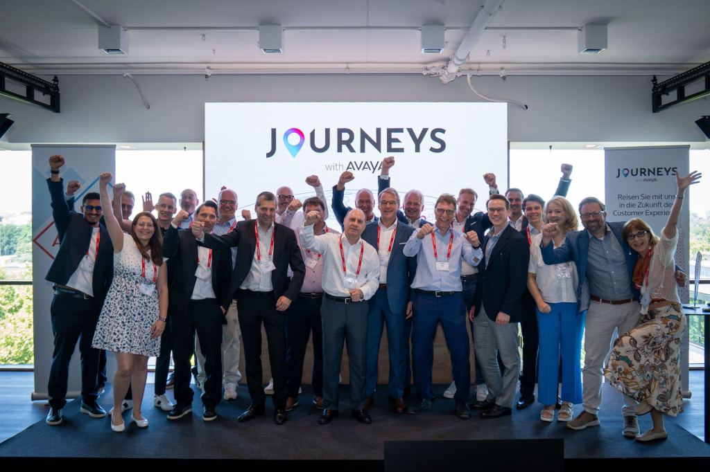 Rund zwanzig Gäste mit Lanyards stehen jubelnd mit gereckten Fäusten vor einer Leinwand mit dem Schriftzug Journeys with Avaya.