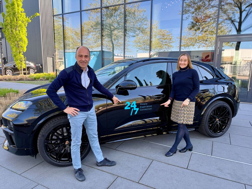 Anja und Daniel Halama stehen links und rechts neben einem schwarzen Porsche Macan und zeigen auf das 24/7-Logo an der Fahrertür.