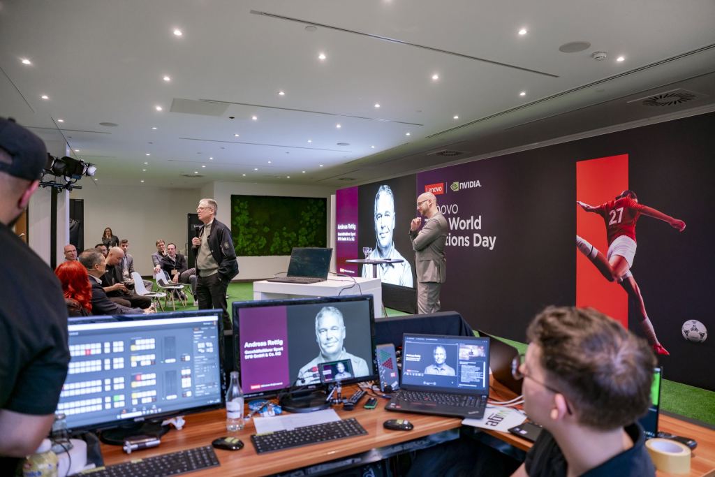 Blick von der Regie auf die Bühne des Lenovo Tech World Solutions Day: zwei Männer im Gespräch vor einer Leinwand, im Vordergrund Mischpulte und Bildschirme mit dem Bild des Redners.