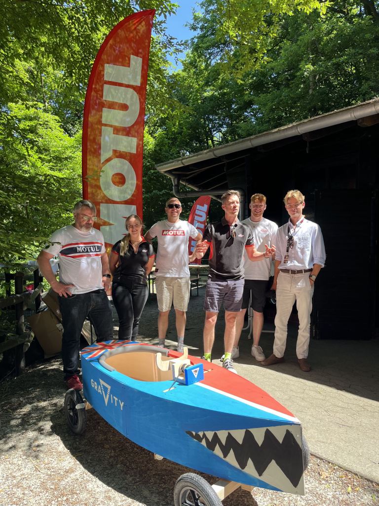 Sechs Personen in Motul-Shirts stehen vor einer Holzhütte hinter einer selbstgebauten Seifenkiste, die als Hai mit Zähnen bemalt ist.
